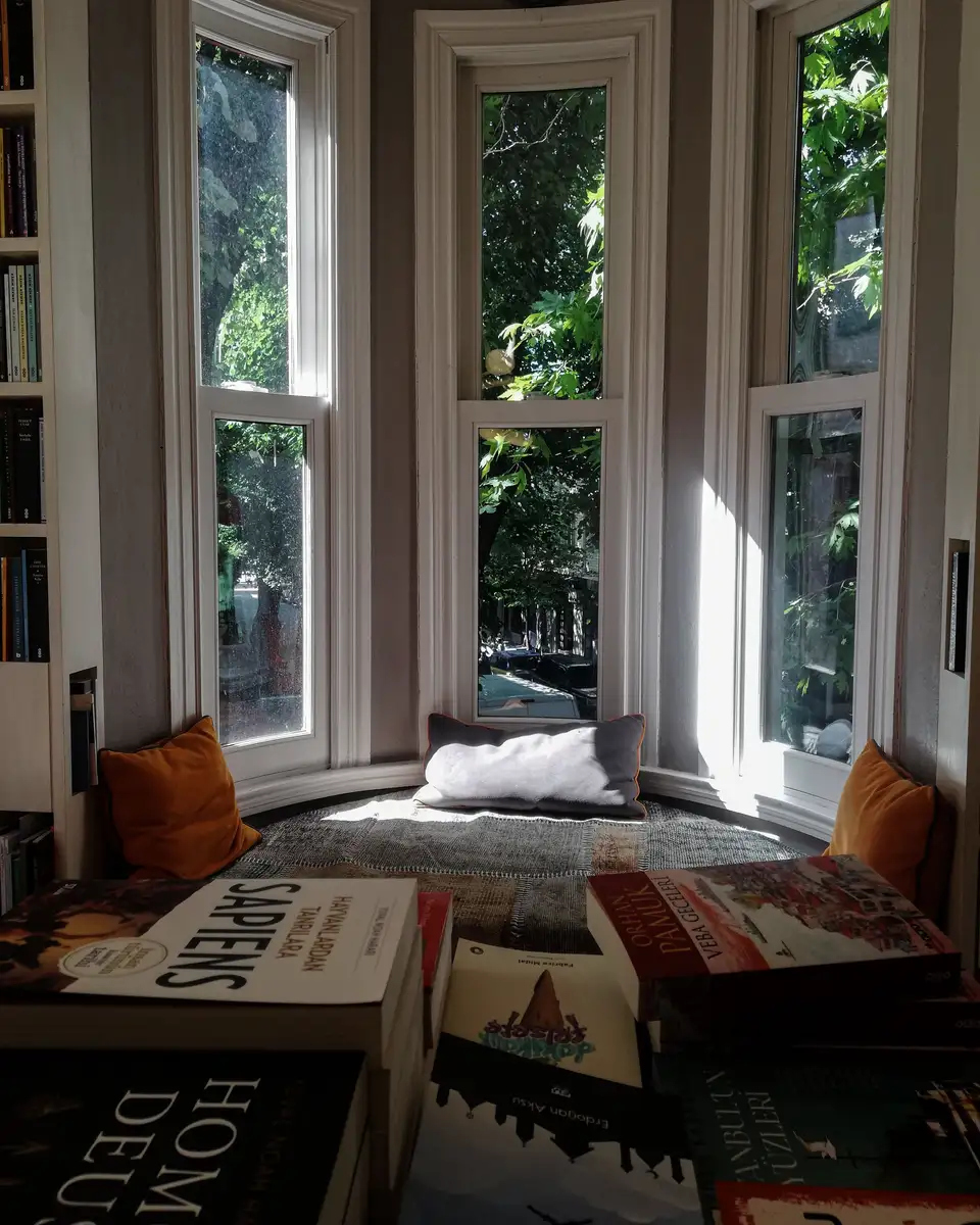 A bay window seat with cushions and books, afternoon light on the glass